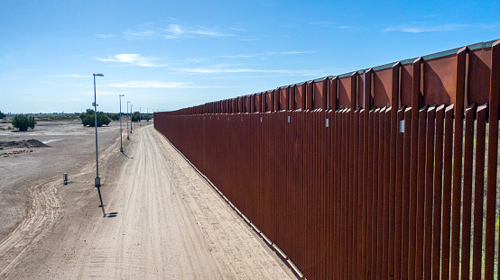 The image shows the imposing US-Mexico border wall stretching across the rugged and arid terrain of the border region. The wall, which is made of concrete and steel, rises up to 30 feet in height. It snakes through the landscape, dividing communities and ecosystems alike. The image also captures the stark contrast between the two sides of the border, with the wall standing as a symbol of the ongoing debate over immigration policy in the United States. The image highlights the complexity and controversy of the US-Mexico border region, where issues of immigration, national security, and human rights intersect. Despite its divisive nature, the wall remains a powerful symbol of the challenges facing both the United States and Mexico in the 21st century.