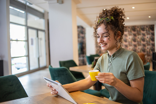 A young beautiful girl is sitting in a coffee shop and looking at her tablet, working online or surfing the internet, browsing social networks, she is dressed in a fashionable youthful style and has curly hair