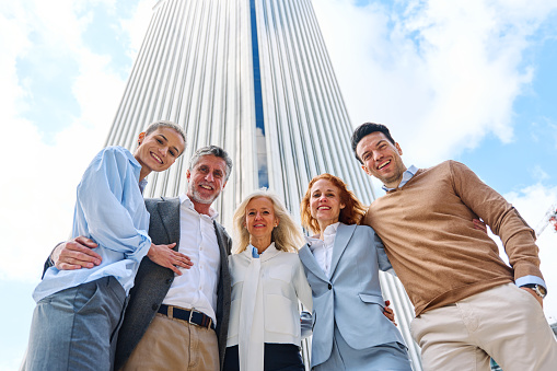 Low angle photo of a group of business people embracing while looking at camera next to a skyscraper