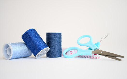Sewing kit consisting of three spools of blue thread in different shades, scissors and measuring tape on a white background. Copy space