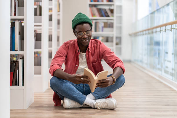 istock Happy African American millennial guy sitting on floor reading book while visiting library 1475957142