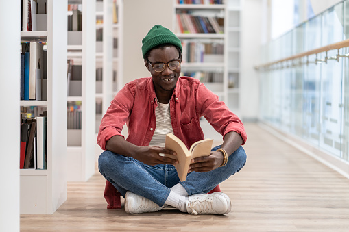 Happy African American millennial guy wearing glasses sitting on floor with book in hands, visiting library, student choosing literature. Smiling black man in bookstore. Reading hobby, self-education