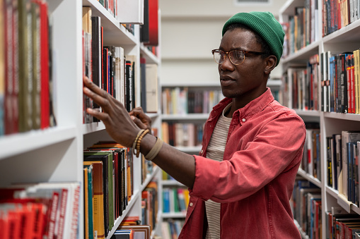 African guy University student wearing glasses standing between bookshelves in library, picking up book from shelf, looking for literature while preparing research project. Education and reading
