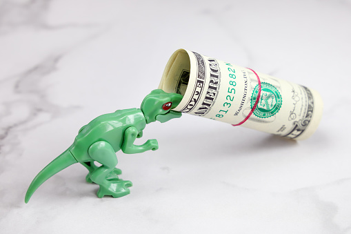 Small green dinosaur holds roll dollar in his teeth, with copy space