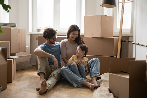 Cheerful couple of parents and cute little daughter kid sitting on heating floor of new home, apartment, resting among stacked cardboard boxes, smiling, laughing, having fun, enjoying relocation