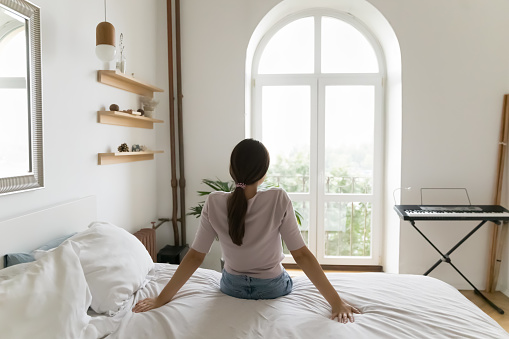 Back view of relaxed young woman sitting on bed with white linen, looking forward at window, enjoying relaxation in bedroom, hotel room, city view, comfort at cozy home