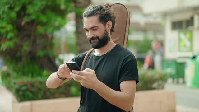 Young hispanic man musician using smartphone holding guitar case at park