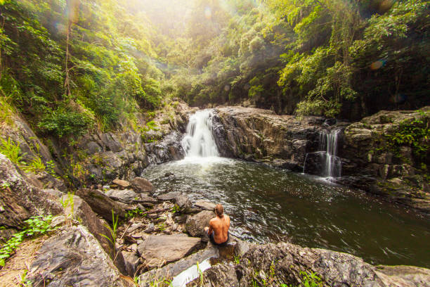 Young man sitting by waterfall flowing over rocks into freshwater swimming hole in lush tropical rainforest scene stock photo