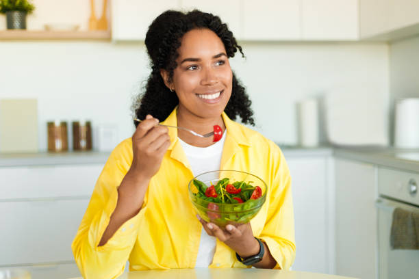 istock Positive black lady eating fresh vegetable salad for dinner, sitting in light kitchen interior at home, free space 1476000574