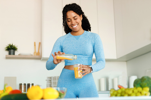 Happy black fit woman having fresh juice for breakfast in the kitchen, pouring beverage from bottle to glass, enjoying healthy eating, free space