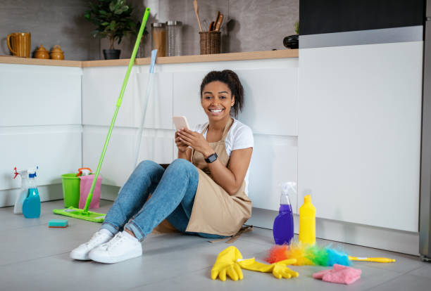 alegre joven negra sentada en el suelo con artículos de limpieza, charlando y navegando por teléfono en la cocina - cleaning fotografías e imágenes de stock