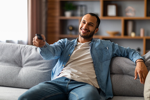 Black Guy Watching TV At Home. Young African American Man With Remote Controller In Hand Resting On Couch In Living Room, Smiling Male Sitting On Sofa And Switching Television Channels, Free Space