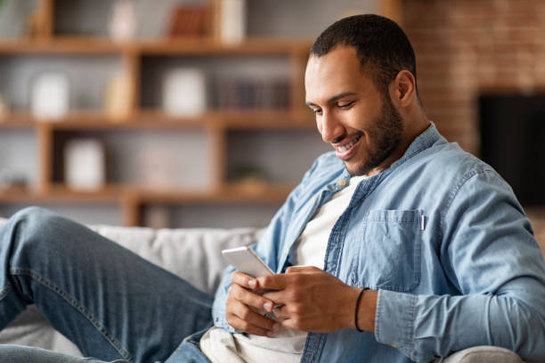 istock Smiling Young Black Man Using Smartphone While Resting On Couch At Home 1476000661