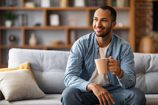 Portrait of handsome african american man resting on couch at home with cup of coffee, young black guy sitting on sofa in living room interior, looking away and enjoying hot drink, copy space