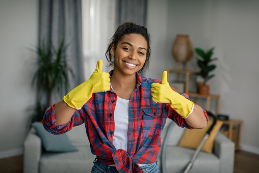 Great work. Happy young black lady in gloves shows thumbs up, enjoys cleaning result in living room interior. Perfectionism at household, cleanliness and hygiene at home alone, lifestyle and emotions