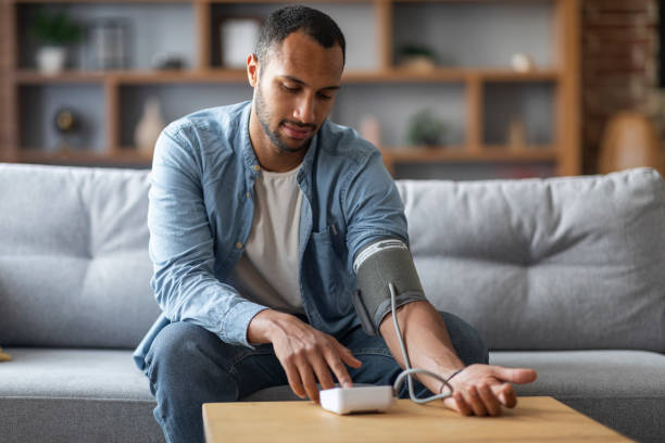 black man sitting on couch, checking blood pressure with upper arm monitor - upper arm fotos imagens e fotografias de stock