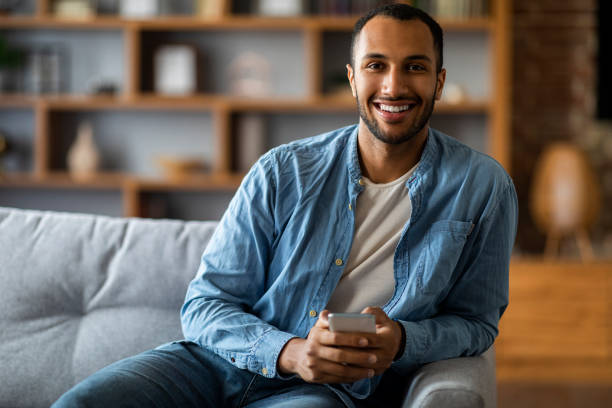 istock Smiling Young Black Man Sitting On Couch With Smartphone In Hands 1476001737