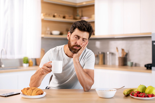 Tired sad sleepy handsome millennial caucasian guy with beard in domestic clothes with cup of coffee sleeps at table, has breakfast in kitchen interior. Waking up early, overwork and time to work
