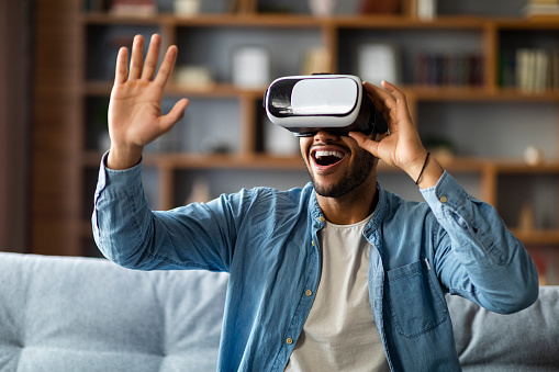 Excited Black Man Using VR Glasses While Relaxing On Couch At Home, Emotional Young African American Guy Experiencing Virtual Reality, Enjoying Modern Technologies For Leisure, Free Space