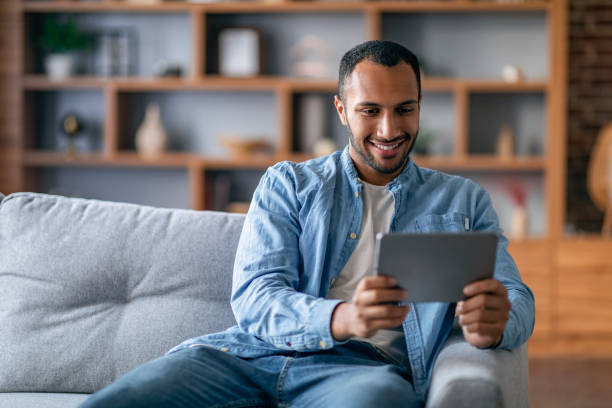istock Cheerful black male watching videos on digital tablet while relaxing on couch 1476002040