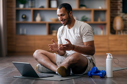 Remote Trainings. Young Black Guy Having Video Call With Sport Coach, Smiling African American Man Talking At Computer Webcam While Exercising On Fitness Mat In Living Room, Copy Space