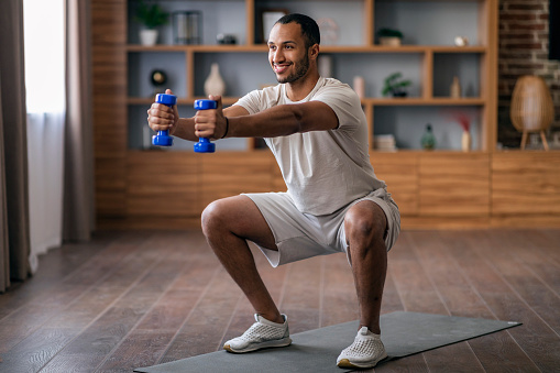 Portrait Of Happy Young Black Man Making Squats And Training With Dumbbells At Home, Smiling Athletic African American Guy Exercising In Living Room Interior, Enjoying Domestic Workout, Copy Space