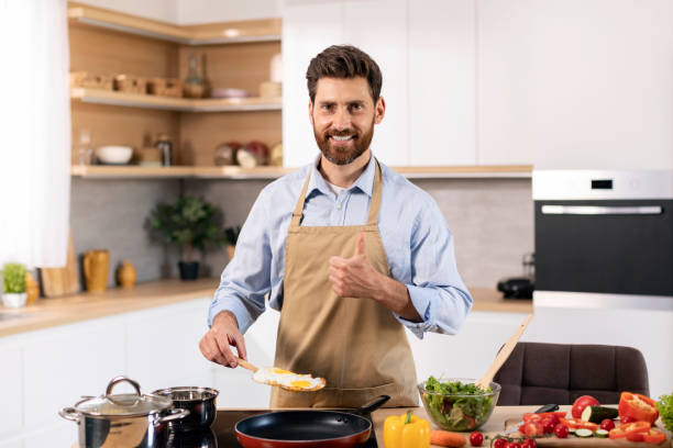 istock Glad millennial caucasian bearded guy chef in apron preparing scrambled eggs and showing thumb up sign 1476002217