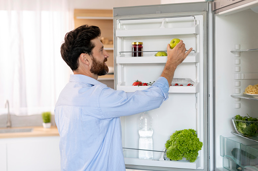 Happy hungry millennial caucasian bearded guy opens refrigerator door and takes green apple for cooking food in kitchen interior, copy space. Diet, vegetarianism, health care and lifestyle at home