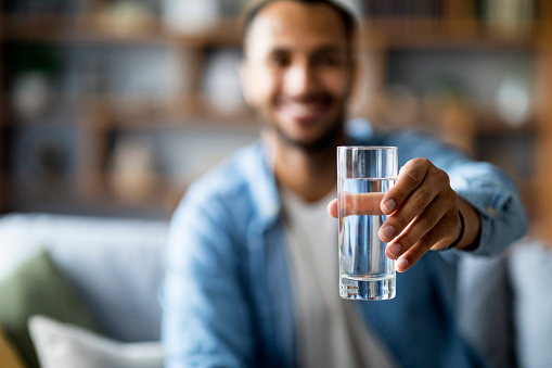 Smiling Young African American Man Giving Glass With Mineral Water At Camera, Happy Handsome Black Male Offering Refreshing Drink, Enjoying Healthy Beverage While Resting On Couch At Home