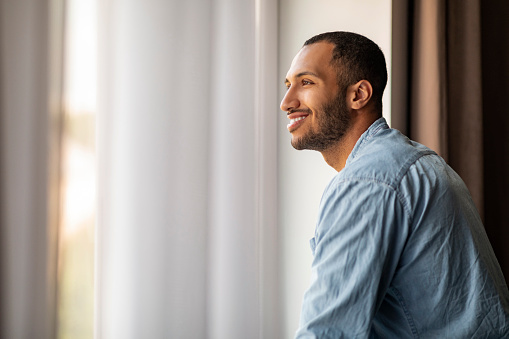Handsome Smiling African American Guy Standing Near Window At Home And Looking Away, Pensive Young Black Man In Casual Clothes Daydreaming Indoors, Having Good Mood, Side View With Copy Space