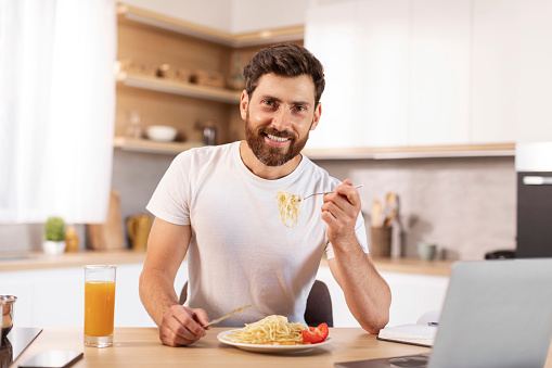 Happy handsome mature caucasian bearded male eating pasta in minimalist kitchen interior with laptop. Good morning, study, work remote, homemade food, lunch and video at home due covid-19 quarantine