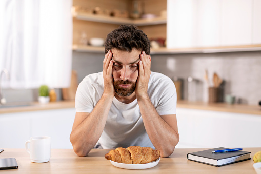 Despaired tired millennial caucasian man with beard holds head with hand, asleep at table with croissant in kitchen interior. Overwork, lack of sleep, emotions early in morning and breakfast at home