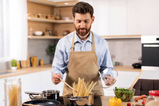 istock Smiling attractive millennial caucasian bearded guy in apron cooks spaghetti at table with vegetables in kitchen 1476002423