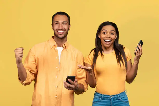 Online win, shopping sale concept. Excited millennial black couple using cellphones, gesturing YES on yellow studio background. Overjoyed young spouses winning lottery or casino bet