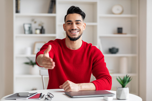 Cheerful friendly attractive young arab businessman with beard in red clothes gives hand and greets at workplace in home office interior. Work and welcomes, customer support and meeting, covid-19