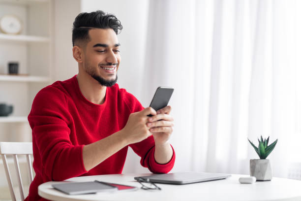 Glad attractive young arabic man with beard in red clothes typing message on smartphone at workplace