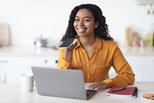 Happy attractive millennnial african amerian woman entrepreneur recording voice message on cell phone with empty screen while working on laptop, sending message to her assistant, kitchen interior