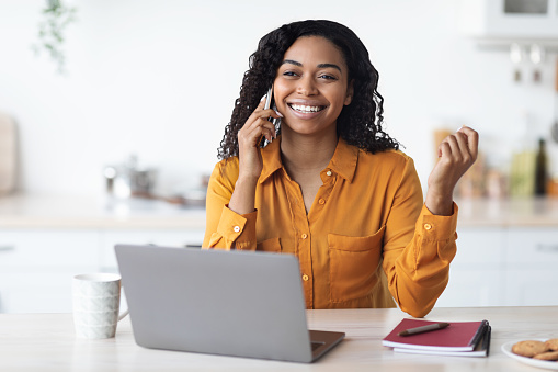 Happy attractive millennnial african amerian woman entrepreneur talking on cell phone while working on laptop, having conversation with her assistant or client, kitchen interior, copy space