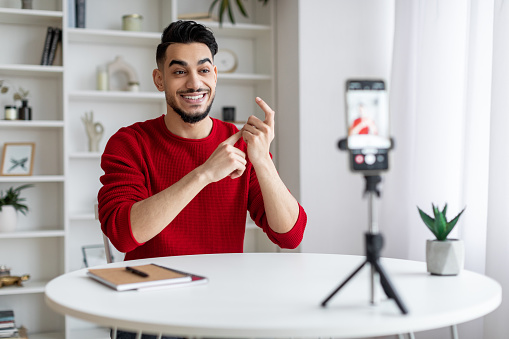 Blogging Concept. Young Arab Guy Recording Video Content For Social Networks, Middle Eastern Male Blogger Using Smartphone On Tripod At Home, Talking And Gesturing At Phone Camera, Copy Space