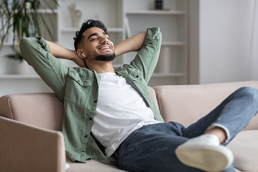 Handsome Arab Guy In Airpods Easrphones Listening Music At Home, Smiling Middle Eastern Man Leaning Back On Couch With Closed Eyes And Hands Behind Head, Enjoying Favorite Playlist, Closeup