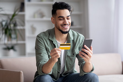 Happy Handsome Middle Eastern Guy Shopping Online With Smartphone And Credit Card While Relaxing On Couch At Home, Young Smiling Arab Man Using E-Commerce For Internet Purchases, Copy Space