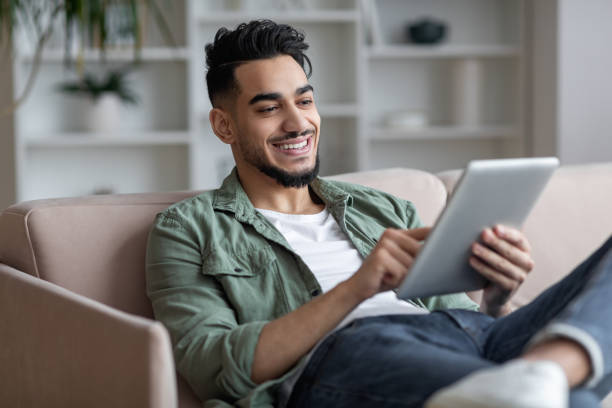 istock Smiling young handsome arab guy relaxing with digital tablet at home 1476004346