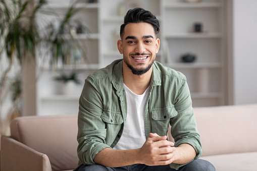 Smiling attractive millennial middle eastern guy blogger with beard looks at camera, sits on sofa in living room interior. Modern blog, study and video call at home due covid-19 outbreak, free space