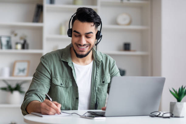 istock Glad young arab guy with beard in headphones works on laptop, watch webinar, make notes 1476004407