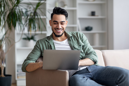 Portrait Of Handsome Young Middle Eastern Guy Using Laptop While Sitting On Couch At Home, Millennial Arab Man Browsing Internet On Computer, Working Remotely Or Shopping Online, Copy Space