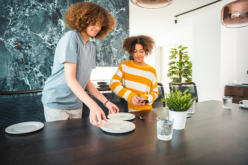 Mixed race teenage girls carrying plates to the table. Setting the table for a family dinner in a beautiful domestic dining room.