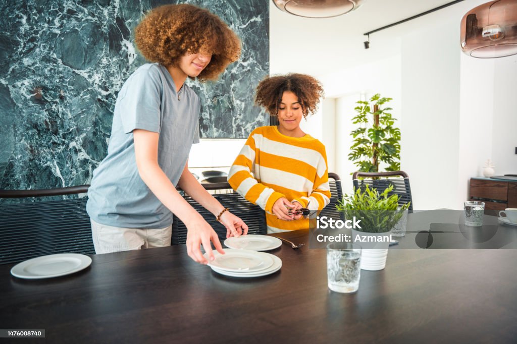 Mixed Race Girls Setting The Table For A Family Dinner Mixed race teenage girls carrying plates to the table. Setting the table for a family dinner in a beautiful domestic dining room. Setting The Table Stock Photo