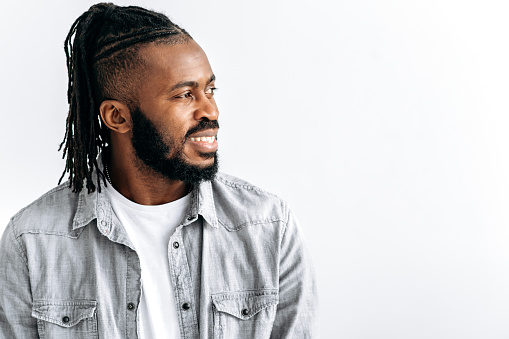 Photo of an attractive black guy, african american man, with dreadlocks, in casual shirt, standing over isolated white background, looking to the side with candid pleasant smile. Copy-space concept