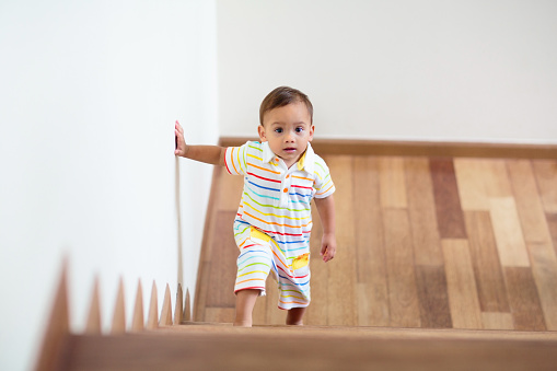 Kid walking stairs in white house. Asian baby boy playing in sunny staircase. Home safety for toddler. Family moving into new home. Child crawling steps of stairway. Foyer or living room interior.