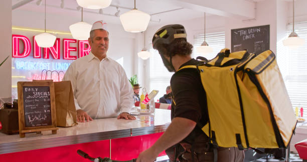 A food delivery worker wearing a cycling helmet picking up an order from the proprietor of a 1950s style diner. He has a large insulated yellow backpack.